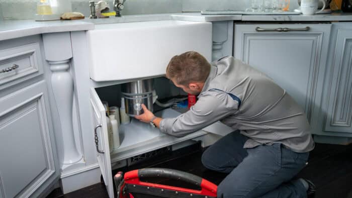 Connor Smith, Owner of ProPipe Plumbing kneels on the floor in a kitchen, repairing a garbage disposal unit under the sink with tools beside him.