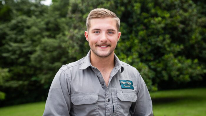 Connor Smith, Owner of ProPipe Plumbing with short hair and a mustache stands outdoors, wearing a gray button-up shirt with a "Pro Pipe plumbing" logo. Green trees and grass are in the background.