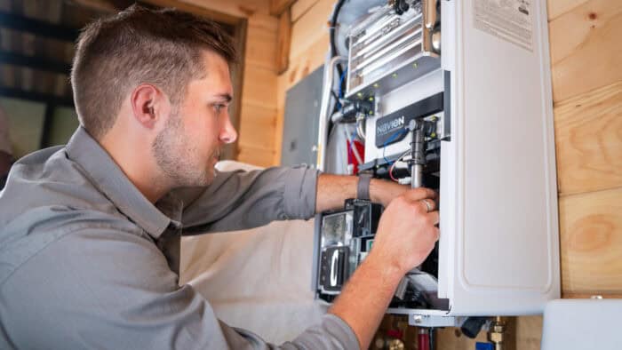 Connor Smith, Owner of ProPipe Plumbing adjusts piping inside an open wall-mounted water heater in a wooden-paneled room.