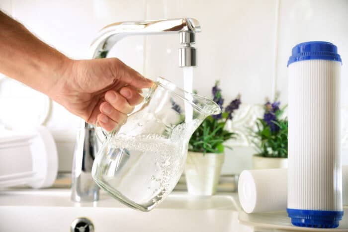 Person filling a glass jug from a tap of purified water with an osmosis system of the home kitchen sink with filters around.